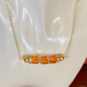 May include: A gold-toned necklace with a delicate chain. The focal point features a row of orange, rectangular beads, interspersed with clear, sparkling accents. The necklace is displayed on a white mannequin.