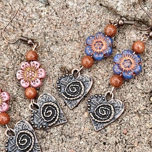 Puede incluir: Dos pares de pendientes con un diseño floral y un dije en forma de corazón. Un par presenta flores rosas con cuentas marrones y el otro par presenta flores azules con cuentas marrones.