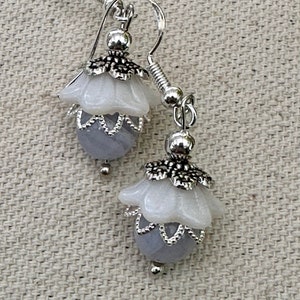 May include: A pair of silver earrings with a white flower design and a light purple bead. The earrings have a delicate, feminine look.