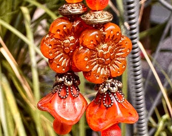 May include: Orange and gold flower-shaped earrings with a bell-shaped dangle. The earrings are made of glass and metal.