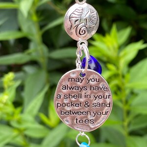 May include: Silver necklace with a charm that says "may you always have a shell in your pocket & sand between your toes". The charm is attached to a silver chain with a blue glass bead and a silver seashell charm.