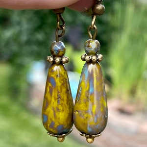 Puede incluir: Un par de pendientes colgantes de tono dorado con cuentas de vidrio en forma de lágrima. Las cuentas son de color amarillo, azul y marrón moteado.