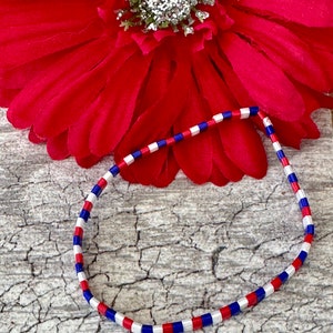 May include: A red, white, and blue beaded bracelet. The bracelet is made of small square beads and is arranged in a repeating pattern of red, white, and blue.