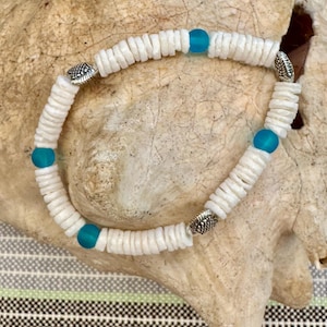 May include: White shell bracelet with turquoise beads and silver-toned accents. The bracelet is made of small, flat, white shells strung together with turquoise-colored beads spaced throughout. The bracelet is displayed on a light brown, textured surface.