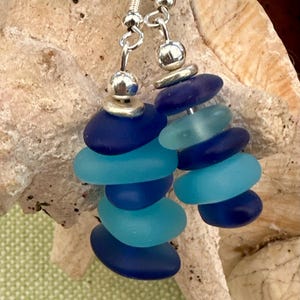 May include: Handmade earrings featuring a cascade of blue and turquoise sea glass pieces. Each earring has a silver-tone metal hook and a decorative silver bead at the top. The sea glass is arranged in a stacked design, creating a unique and eye-catching accessory.