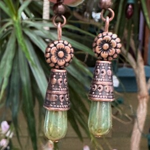 May include: A pair of copper dangle earrings with green glass teardrop beads. The earrings feature a floral design with intricate details.