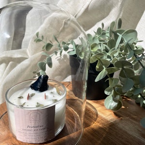 May include: A clear glass candle holder with a white candle and a black stone on top. The candle is labeled "Protection" and contains eucalyptus, peppermint, and lavender. The candle sits on a wooden surface with a glass dome and a small potted plant.