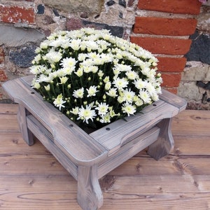 May include: A white wooden planter box with a square shape and four legs. The planter is filled with a white flower arrangement. The planter is sitting on a wooden surface.