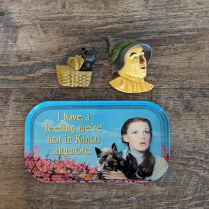 May include: A selection of Wizard of Oz themed items. Features a small black dog in a basket, a Scarecrow head, and a blue tray with the quote "I have a feeling we're not in Kansas anymore" and a picture of Dorothy and Toto.