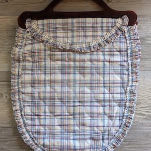 May include: A vintage quilted bag with a dark brown handle. The bag features a white background with a plaid pattern in red, blue, and yellow. The edges are trimmed with a ruffled fabric.