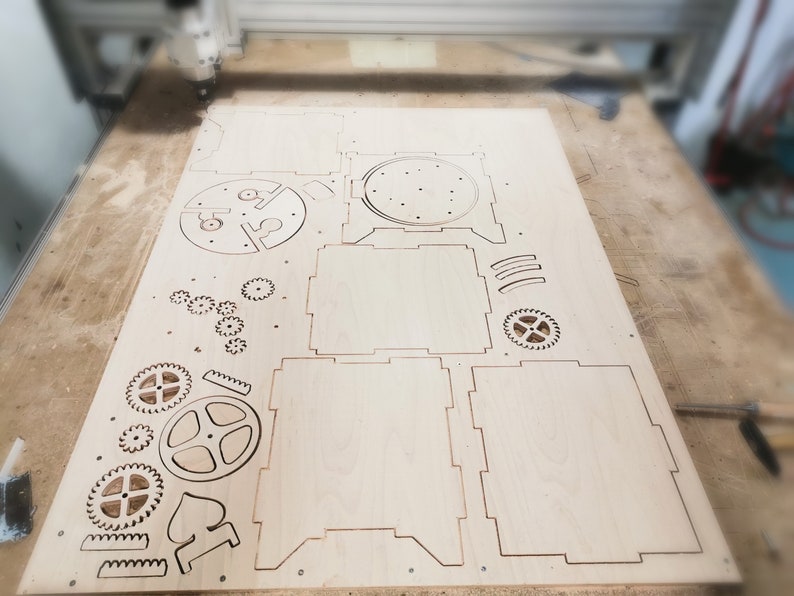 May include: A sheet of plywood with various shapes cut out of it, including gears, a box, and a circle with holes. The plywood is light brown in color and has a smooth surface.