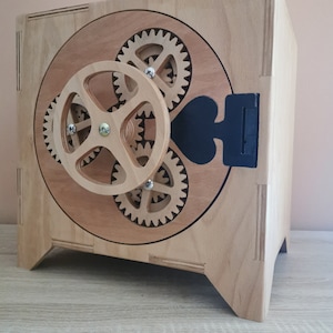 May include: A wooden box with a round, open front featuring a gear mechanism. The box is made of light wood with a natural finish. The gears are made of a darker wood and are visible through the open front. The box has a black handle on the side.