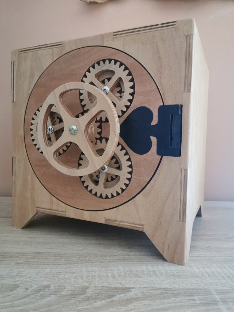 May include: A wooden box with a round, open front featuring a gear mechanism. The box is made of light wood with a natural finish. The gears are made of a darker wood and are visible through the open front. The box has a black handle on the side.