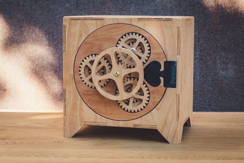 May include: A wooden box with a round, open door featuring a gear mechanism. The box is made of light brown wood and has a black metal handle.