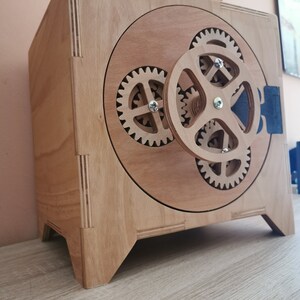 May include: A wooden box with a round, open front that features a series of interlocking gears. The box is painted in a light wood color and has a dark brown handle.