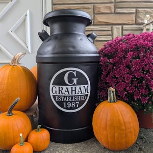 May include: A black metal milk can with the name "GRAHAM ESTABLISHED 1987" in a white circular design. The can is surrounded by orange pumpkins of various sizes and a pot of purple flowers, creating a fall-themed display.