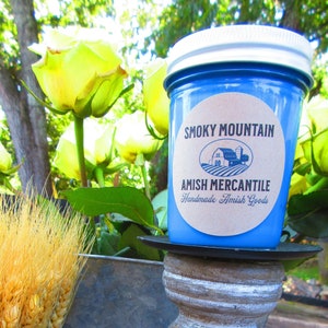 Peut inclure: Un pot en verre bleu avec un couvercle blanc et une étiquette brune qui dit "Smoky Mountain Amish Mercantile Handmade Amish Goods". Le pot est posé sur un piédestal en bois avec un seau en métal gris en arrière-plan.