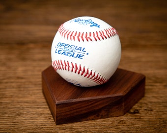 May include: A white baseball with red stitching and blue text, labeled "OFFICIAL LEAGUE" and "Rawlings." The baseball rests on a dark brown, hexagonal wooden base, set against a wooden background.