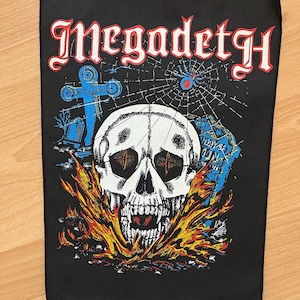 May include: A rectangular black fabric patch featuring the white text Megadeth at the top. The graphic depicts a white skull with red eyes surrounded by orange and yellow flames, a blue cross, a spider web, and a tombstone on a black background.