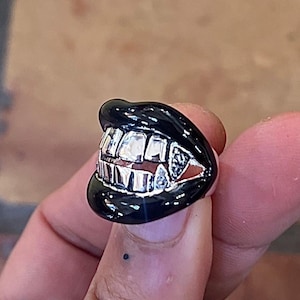 Op de afbeelding: Een zwarte ring met een zilveren grillontwerp. De ring is gevormd als een mond met tanden.