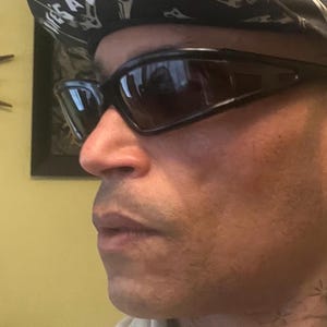 Könnte beinhalten: Schwarze Sonnenbrille mit dunklen Gläsern. Die Person trägt ein schwarz-weiß gemustertes Bandana. Der Hintergrund umfasst eine dekorative Uhr und ein gerahmtes Bild.