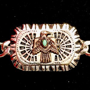 Op de afbeelding: Zilveren armband met een rechthoekige bedel met een gestileerde adelaar met een turquoise steen in het midden. De bedel heeft een gedetailleerd ontwerp met concentrische cirkels en een getextureerde rand.