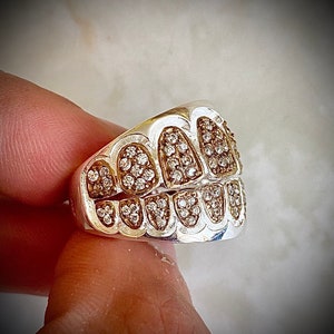 Könnte beinhalten: Ein silberner Ring mit einem diamantbesetzten Design, das einer Zahnreihe ähnelt. Der Ring sitzt an einem Finger.