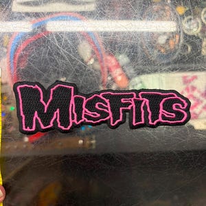 Può includere: Una toppa ricamata nera con bordo rosa che presenta la parola MISFITS in un carattere stilizzato e frastagliato. La toppa è posizionata accanto a un metro a nastro giallo, che mostra una larghezza di circa 11,5 cm e un'altezza di 3,8 cm.