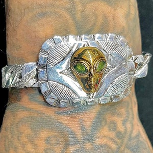 Puede incluir: Una pulsera de plata con un dije de cabeza de extraterrestre de oro. El extraterrestre tiene ojos verdes y una cara detallada.