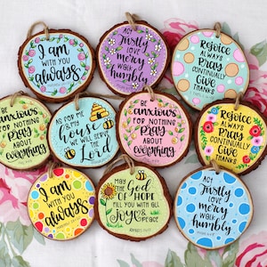 May include: Ten round wood slices with painted inspirational quotes and bible verses. The wood slices are decorated with flowers, bees, and polka dots. The quotes include "I am with you always", "Act justly, love mercy, walk humbly", "Be anxious for nothing, pray about everything", "Rejoice always, pray continually, give thanks", and "May the God of hope fill you with all joy and peace".