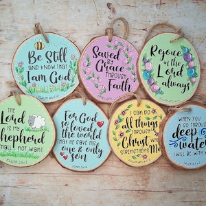May include: Set of 8 round wooden ornaments with painted floral designs and inspirational bible verses. The ornaments are painted in various colours, including pink, blue, yellow, and green. Each ornament features a different verse from the bible, such as "Be Still and Know That I Am God", "Saved by Grace Through Faith", "Rejoice in the Lord Always", "The Lord is My Shepherd, I Shall Not Want", "For God So Loved the World That He Gave His One and Only Son", "I Can Do All Things Through Christ Who Strengthens Me", "When You Go Through Deep Waters I Will Be With You".