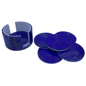 May include: A set of six round, dark blue coasters with a white trim, alongside a matching holder. The coasters and holder are made of a stone-like material. The holder is a curved shape with a flat base.
