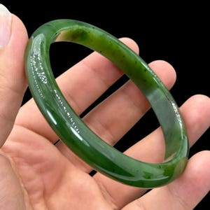 Puede incluir: Un brazalete rígido liso y pulido, elaborado en jade verde oscuro sólido. El accesorio circular presenta una calidad translúcida con sutiles inclusiones naturales y variaciones en la intensidad del color a lo largo de toda la superficie redondeada y continua.