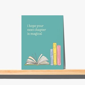 May include: A teal greeting card featuring a watercolor illustration of an open book and three standing books in blue, yellow, and pink. The text "i hope your next chapter is magical" is printed in white on the upper left side of the card.