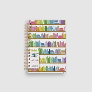 May include: A white spiral bound notebook with a colorful bookshelf design featuring rows of colorful books. The notebook cover has the text "emmy + olly" and "notebook" printed on it.