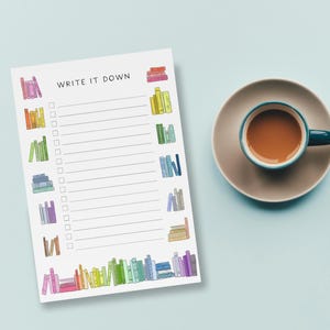 May include: A white notepad with the words "WRITE IT DOWN" and a series of checkboxes. The notepad is decorated with colorful illustrations of stacked books. A teal mug filled with a brown liquid sits on a saucer to the right.