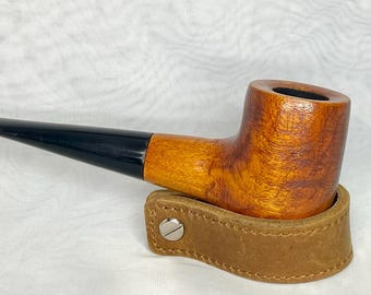 May include: A wooden tobacco pipe with a black stem and a brown, textured bowl. The pipe rests on a brown leather holder with a silver button. The pipe has a classic, curved design.
