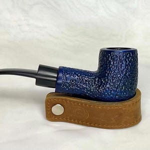 May include: A blue tobacco pipe with a textured surface, resting on a brown leather holder. The pipe has a black stem and a curved design. The leather holder has a silver button closure.