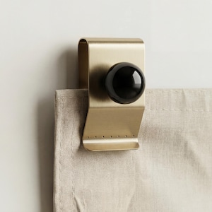 May include: A brass-colored curtain clip with a black, spherical knob. The clip is attached to a beige curtain. The clip has a curved top and a series of small holes along the bottom edge.