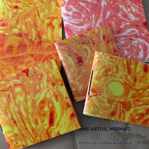 May include: A collection of notebooks with covers featuring a vibrant, swirled pattern in shades of yellow, orange, and red. Some notebooks are open, revealing the interior pages. The text "THIS ARTFUL MERMAID" and social media handles are visible.