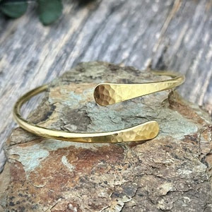 May include: A gold hammered bangle bracelet with a simple, open design. The bracelet is made of thick, textured metal and has a slightly curved shape.
