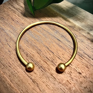 May include: A gold bangle bracelet with two round ends. The bracelet is on a wooden surface.