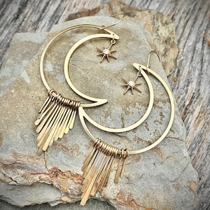 May include: Gold tone earrings with a crescent moon design and a dangling star charm. The earrings have a fringe of thin metal strands.