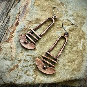 May include: A pair of copper earrings with a geometric design. The earrings are long and dangle, with a curved shape at the bottom. The earrings are made of hammered copper, giving them a rustic look.