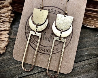 May include: Gold geometric earrings with a crescent moon, rectangle, and a half-circle shape. The earrings are on a brown card with the text "Made with Love Glow Studio."