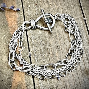 May include: A silver chain bracelet with a toggle clasp. The bracelet is made of three different chain styles, including a curb chain, a box chain, and a cable chain.