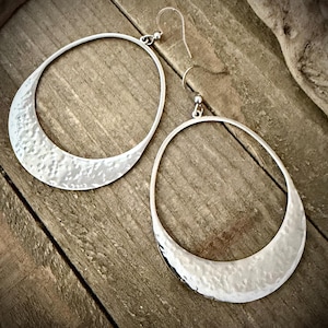 May include: A pair of silver-toned earrings featuring large, open teardrop-shaped hoops. The lower portion of each hoop has a textured, hammered finish, while the upper section remains smooth and thin, attached to standard silver-toned ear wires.