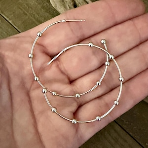 May include: A pair of silver hoop earrings featuring a thin circular wire design accented with small, evenly spaced silver beads along the circumference. The earrings have a polished metallic finish and include a standard post closure mechanism.