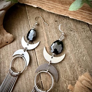 May include: A pair of silver earrings with a celestial design. Each earring features a faceted black bead, a crescent moon, a circular ring, and multiple dangling silver bars. The earrings are displayed on a wooden surface.