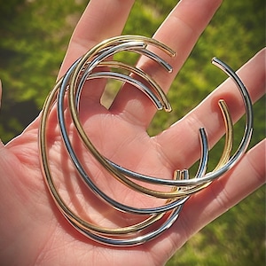 May include: A collection of open-ended bangles in gold and silver tones. The bracelets are displayed in a hand, showcasing their simple, circular design. The metal jewelry reflects light, creating a bright and elegant appearance. The background is a blurred green.
